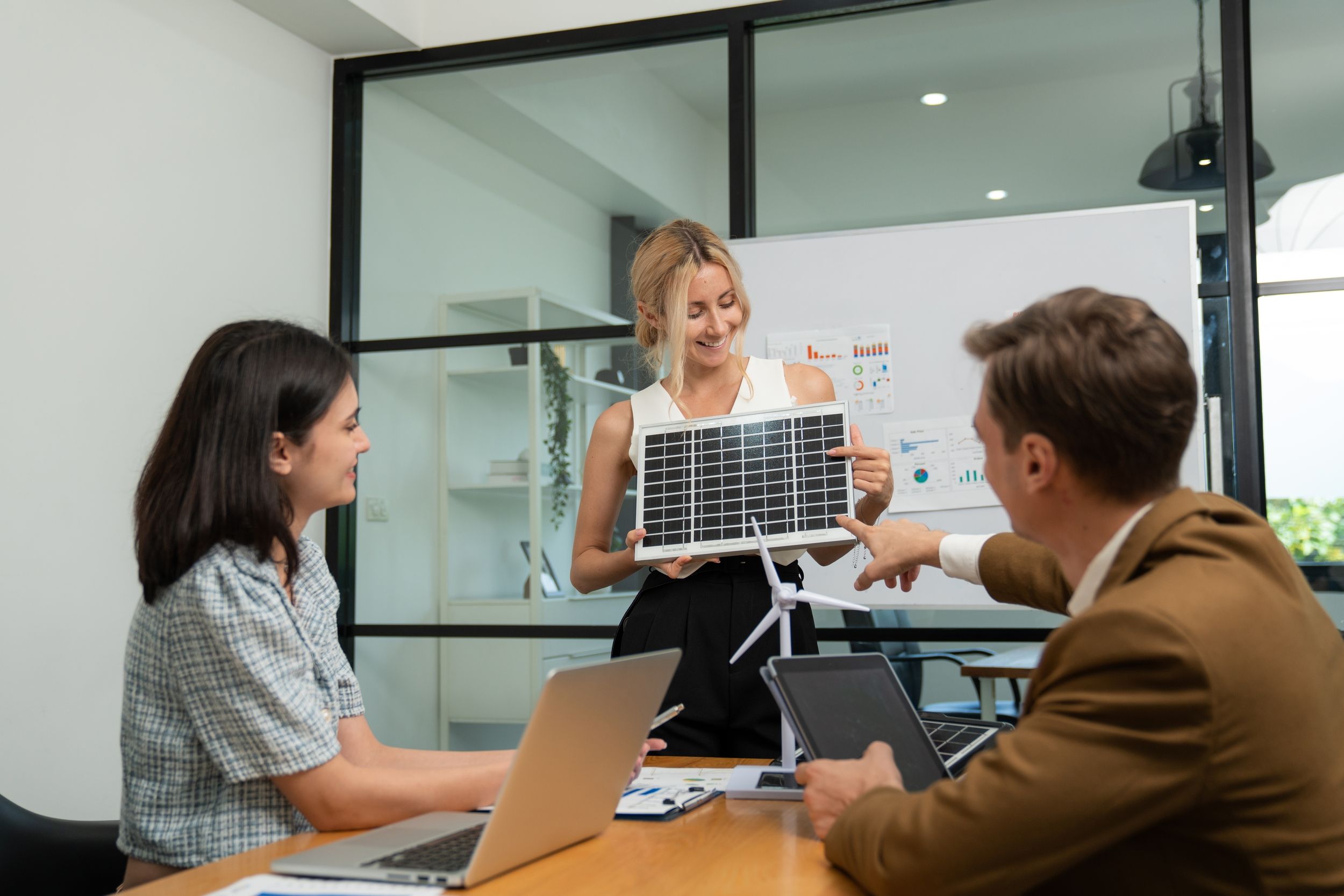 eine Energieberaterin hält eine Photovoltaik-Atrappe in der Hand und zeigt es zwei Kunden